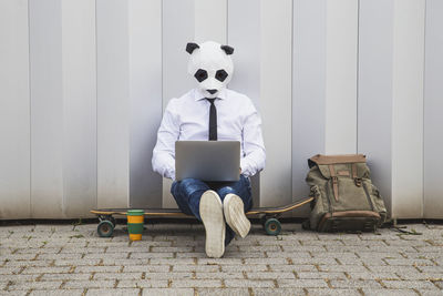 Businessman wearing panda mask and laptop while sitting on skateboard against gray wall.