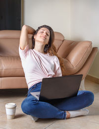 Relaxed young women sitting on the floor with laptop and coffe to go near sofa 