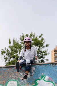 Full body of cheerful young african american female wearing white t shirt with helmet and safety pads with skateboard sitting on skate ramp in park