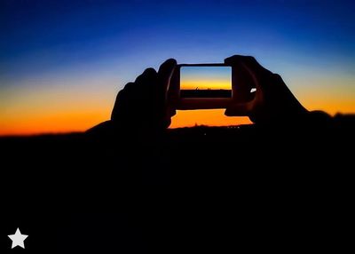 Silhouette hand holding smart phone against sky during sunset
