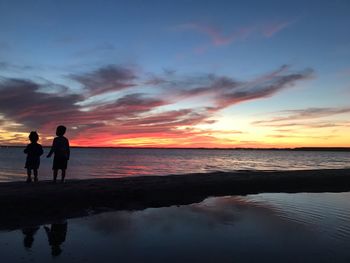 Silhouette siblings standing at beach against sky during sunset