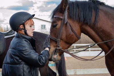 Red hair young woman in equestrian helmet pets horse