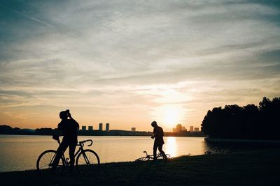 Silhouette people with bicycles at lakeshore against sky during sunset