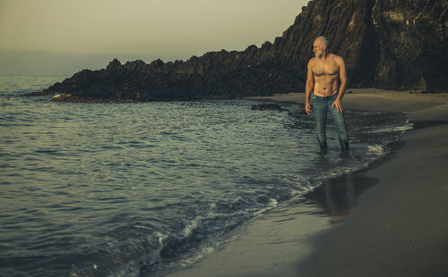Shirtless adult man in jean walking on beach during sunrise