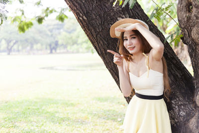 Portrait of beautiful young woman wearing hat while standing by tree trunk
