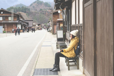 Side view of young woman sitting on bench by house