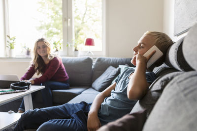 Male teenager talking on phone while smiling sister sitting on sofa at home