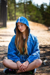 A young girl poses in a denim jacket with a hood against the background of a forest sitting