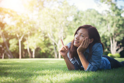 Thoughtful young woman pointing upwards while lying on grass at park
