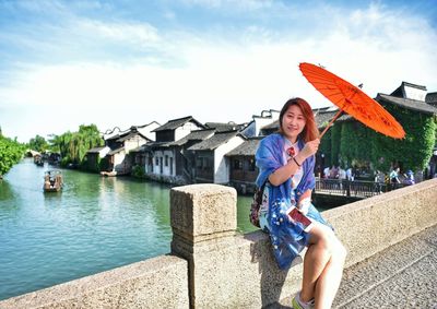 Portrait of woman with umbrella sitting on railing against cloudy sky