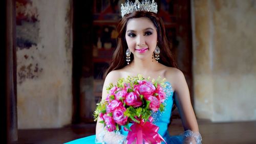 Bride wearing crown while holding pink rose bouquet by wall