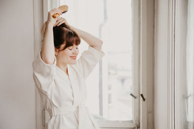 Young woman looking away while standing against white wall at home