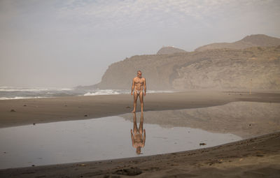 Man with swimwear standing on beach with reflection on water against sky