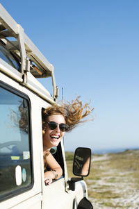 Happy woman in sunglasses peeking through car window during sunny day