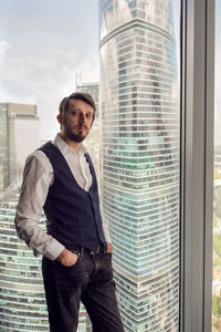 Thoughtful businessman with beard stands next to  window in office backdrop of skyscrapers in moscow