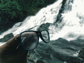 Cropped hand holding eyeglasses against waterfall