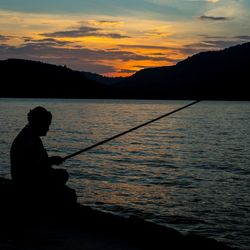 Silhouette man fishing in lake against sunset sky