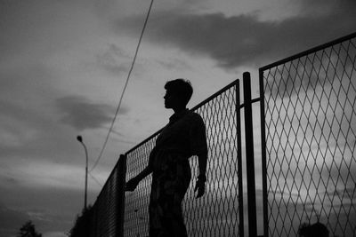 Silhouette man standing by fence against sky