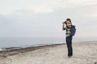 Full length of woman carrying backpack photographing sea against sky