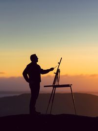 Silhouette man painting while standing against sky during sunset