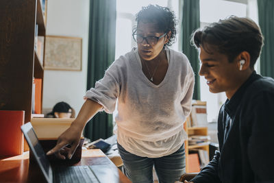 Teacher pointing at laptop while teaching smiling male student in library