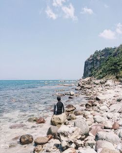 Rear view of man sitting on rock at shore against sky