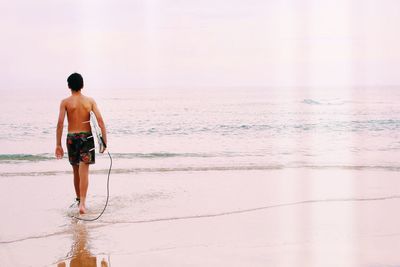 Rear view of shirtless boy with surfboard walking at beach against sky