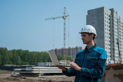 A male builder in a helmet stands against the background of a construction site with a laptop in his