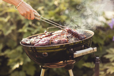 Cropped hand of woman grilling meat with serving tongs in back yard on sunny day