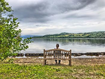 Rear view of woman sitting on bench by lake against sky