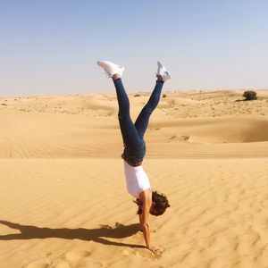 Young woman doing handstand on sand at desert against clear sky