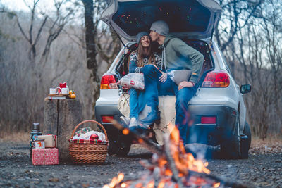 Couple embracing while sitting at car trunk by bonfire at forest