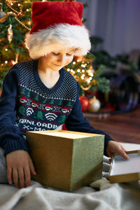 A happy boy in a santa claus hat opens a box with a christmas present next to a christmas tree