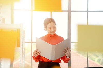 Woman reading file while standing in office seen through glass window