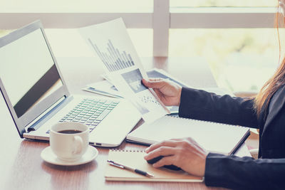 Midsection of woman with coffee analyzing graphs by laptop on table