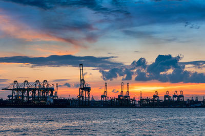 Cranes at harbor against sky during sunset