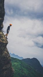 Low angle view of young man rock climbing against cloudy sky