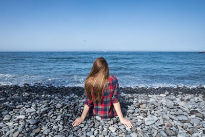 Rear view of woman sitting on rocks at beach against sky