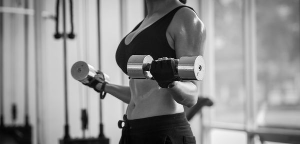 Midsection of woman holding dumbbells and exercising at gym