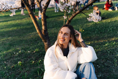 Smiling cheerful woman in white down jacket and jeans sitting on grass in city park in spring