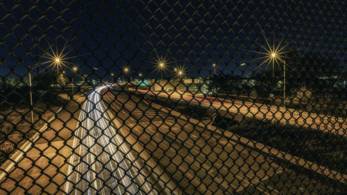 Chainlink fence by illuminated street against sky at night