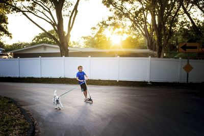 Full length of playful boy with dog riding push scooter on road during sunset