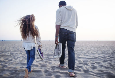 Rear view of father and daughter carrying shoes while walking at beach against clear sky