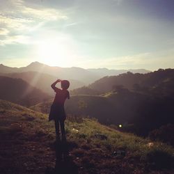 Rear view of woman shielding eyes while standing at mountain against sky during sunset