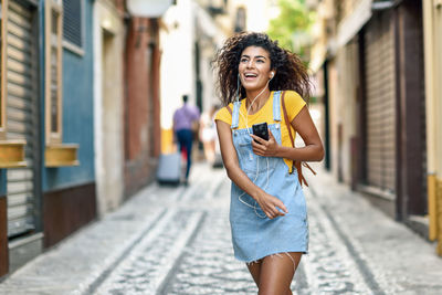 Smiling young woman listening to music while walking at alley in city