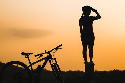 Silhouette of woman standing on wooden stump by bicycle against sky during sunset