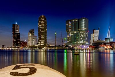 Illuminated buildings by river against blue sky at night