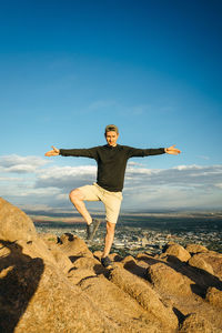 Full length of man standing on one leg over rock against sky