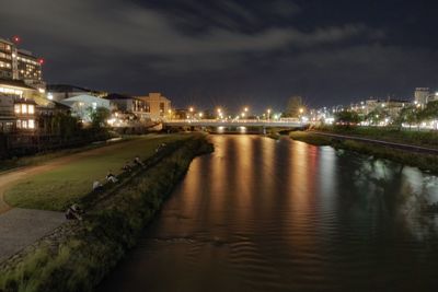 Illuminated street by river and buildings against sky at night