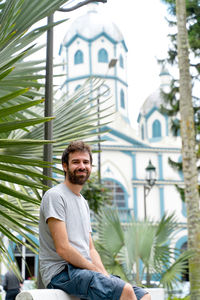 Portrait of young man sitting against buildings in city